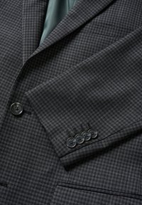 Grå rutiga kavaj med ett texturerat tyg, med slag och detaljer med fem knappar vid ärmarna. Foder synligt i grönt.