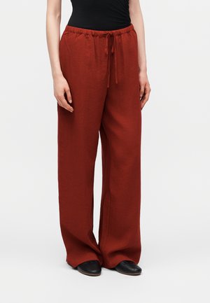 Kobieta ubrana w luźne, rdzawe spodnie ze sznurkiem i czarne buty, stoi na jednolitym jasnym tle.