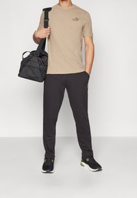 Beige kortärmad atletisk tröja med strukturerad mönster, svarta träningsoverallsbyxor med logotyp, och svarta sportskor med gröna detaljer. Bär en svart väska.