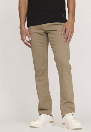 Pantalon beige à coupe droite en mélange de coton, avec un design classique à cinq poches. Associé à des baskets blanches, porté avec une chemise noire.