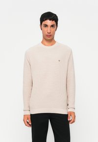 Homme aux cheveux courts et foncés, portant un pull en maille beige clair et un pantalon noir, se tenant devant un fond blanc uni.
