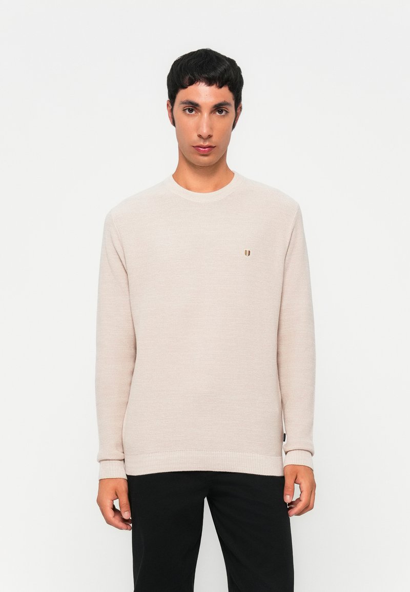 Homme aux cheveux courts et foncés, portant un pull en maille beige clair et un pantalon noir, se tenant devant un fond blanc uni.