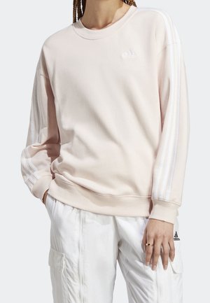 Person iført lyserød Adidas-sweatshirt med hvide striber på ærmerne og hvide cargobukser, stående med den ene hånd i lommen.