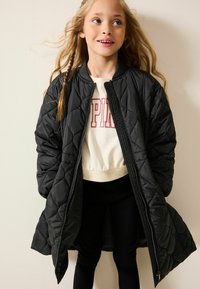 Jachetă bomber căptușită, de culoare neagră, cu un design matlasat, închidere cu zip și manșete ribbate, purtată peste un hanorac scurt alb și colanți negri.