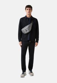 Grå polstret crossbody taske bæret over en navy jakke og sorte bukser, med en glat tekstur og justerbar rem, komplementeret af grå sneakers.