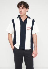 Polo à manches courtes rayé bleu marine et blanc avec un col en V, arborant un logo brodé rouge sur la poitrine. Tissu en coton lisse.