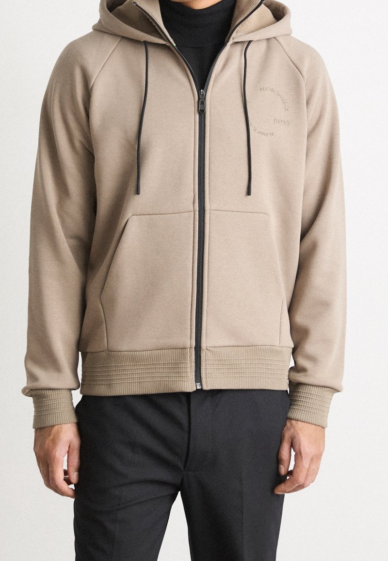 Homme portant un sweat à capuche beige zippé avec des cordons noirs, par-dessus un t-shirt noir et un pantalon noir, debout devant un fond uni.