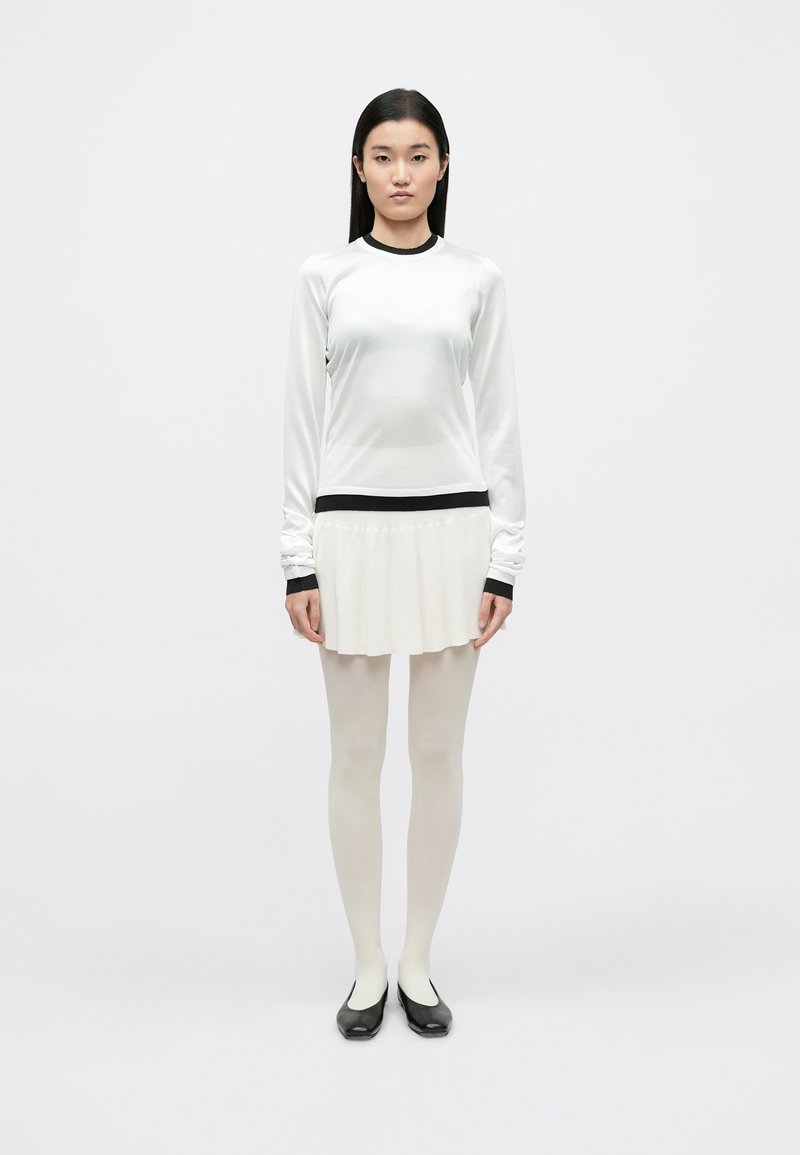 Haut blanc à manches longues avec bordure noire, assorti à une jupe plissée crème. Le mannequin porte des collants blancs transparents et des chaussures plates noires. Design simple, texture lisse.