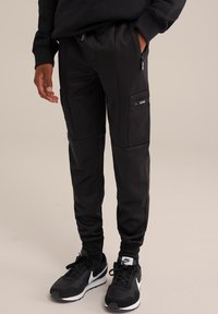 Pantaloni sportivi neri realizzati in tessuto morbido con tasche con zip, design affusolato e polsini a coste. Indossati con sneaker nere Nike.