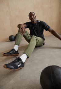 Man in zwart shirt en olijfgroene broek die op de vloer in de sportschool zit met medicine balls in de buurt, lachend naar de camera.