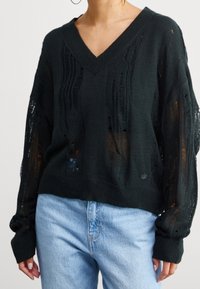 Černý pletený svetr s výstřihem do V, dlouhými rukávy a otvorovou texturou. Obsahuje žebrované manžety a malé logo na lemu.