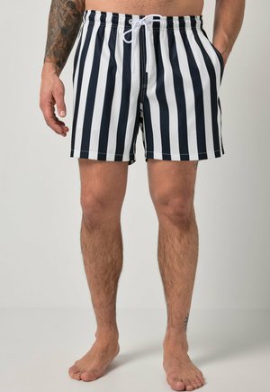 Man draagt zwart-witte verticaal gestreepte zwemshort, staat op blote voeten met zichtbare getatoeëerde linkerarm, handen in de zakken.