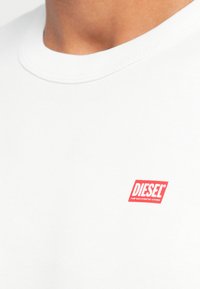 Biała bawełniana koszulka z okrągłym dekoltem, z czerwonym prostokątnym logo "DIESEL" na górnej lewej stronie. Gładka faktura, minimalistyczny design.