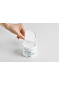 Witte cilindrische container gelabeld "Moisture Up Pad" met een gestructureerd deksel. Een hand haalt een ronde, gestructureerde pad eruit.