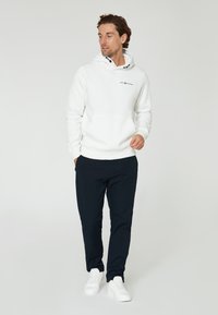 Sudadera blanca con bolsillo frontal, capucha con cordón y logo. Combinada con pantalones azul marino y zapatillas blancas. Estilo suave y casual.