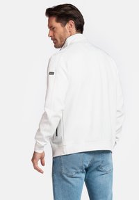 Witte zip-up jack met een staande kraag, geribde manchetten en een zwart logo-patch op de linker mouw. Gekozen bij een blauwe jeans.