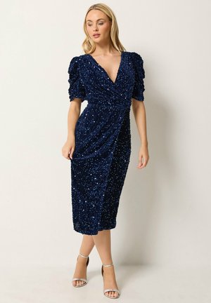 Donna che indossa un vestito a portafoglio blu navy con paillettes, maniche a sbuffo e sandali argento con tacco alto, in piedi su uno sfondo chiaro e uniforme.