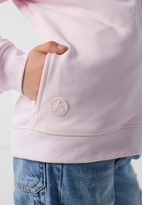 Felpa rosa chiaro con tasca laterale e texture morbida. Presenta un piccolo logo in rilievo dello stesso rosa sulla tasca. Jeans in denim blu visibili.