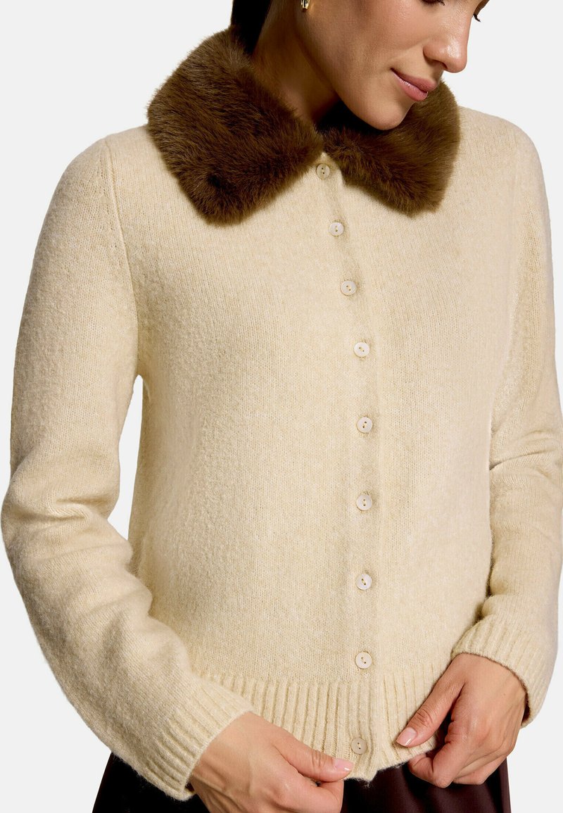 Femme portant un cardigan crème boutonné avec un col en fausse fourrure marron foncé, ajustant le bouton du bas avec ses mains.
