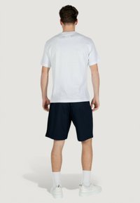 T-shirt bianca in cotone con maniche corte, taglio dritto e etichetta sul retro, abbinata a pantaloni corti blu scuro e sneakers bianche.