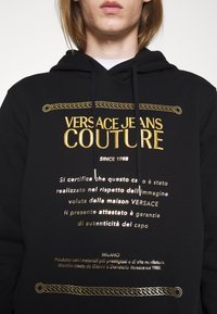 Osoba v černé kapuci s nápisem v zlaté a bílé barvě Versace Jeans Couture a dekorativními řetězovými vzory na přední straně.