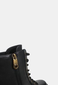 Botín de cuero negro con tirador dorado en el cierre y cordones negros pasados por ojales dorados sobre un fondo claro y liso.