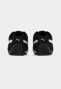 Ζευγάρι από μαύρα και λευκά αθλητικά παπούτσια Puma, εμφανιζόμενα από την πίσω πλευρά, με επενδεδυμένους γιακάδες και χρυσά λογότυπα Puma πάνω από υφασμένα μαύρα πέλματα.