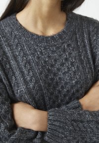 Pull en maille épaisse, gris foncé, avec un motif en torsades, présentant un col rond et des poignets larges côtelés. Sa texture douce et sa coupe décontractée le rendent très agréable à porter.