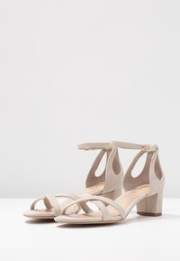 Beige semišové sandály na blokovém podpatku s řemínkem kolem kotníku a dvěma předními řemínky, vystavené na bílé ploše na světlém jednobarevném pozadí.
