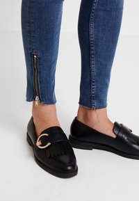 Mocasines de cuero negro con un acento dorado y detalles de flecos, combinados con jeans de denim azul oscuro que tienen un tobillo con cremallera.