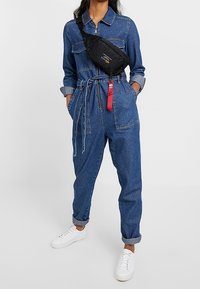 Person i uppvikt denimoverall med knytbälte, vita sneakers och svart crossbody-väska med röd tagg, står med händerna i fickorna.