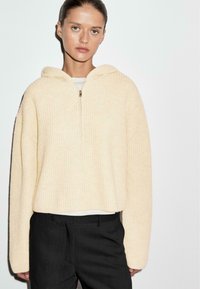 Felpa corta color crema realizzata in materiale a costine, con zip a metà e cappuccio, abbinata a pantaloni neri aderenti.