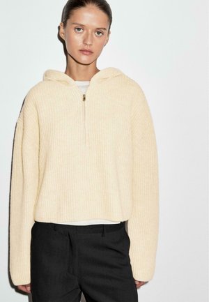 Cremfarbener, cropped Hoodie aus Rippmaterial mit halbem Reißverschluss und Kapuze, kombiniert mit eng geschnittenen schwarzen Hosen.