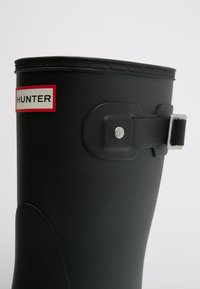 Czarny kalosz Hunter z czerwoną i białą naszywką z logo oraz detalem na bocznej klamrze na jednolitym szarym tle.