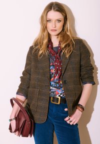 Femme aux longs cheveux blonds portant un blazer à carreaux marron, un t-shirt graphique, un pantalon bleu, une écharpe rouge, tenant un sac à main bordeaux devant un arrière-plan clair.