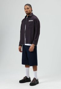 Overhemd met knoopsluiting van donker geruit stof met de tekst "FREEDOM", gecombineerd met knielange denim shorts en stevige zwarte sneakers met roze veters.