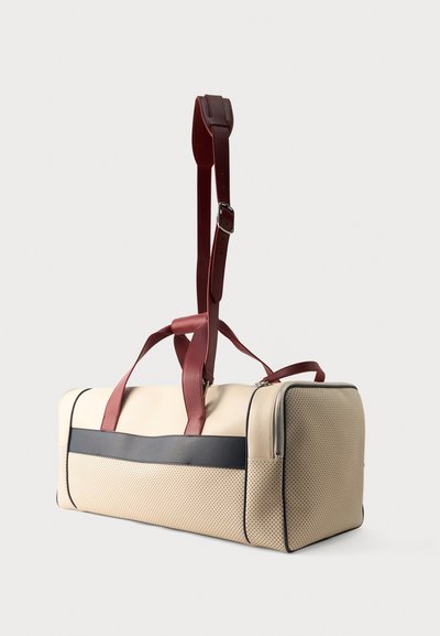 Μπεζ ορθογώνια τσάντα duffel με διάτρητη υφή, μαύρη οριζόντια ρίγα και ρυθμιζόμενους σκούρους κόκκινους ιμάντες ώμου και χειρός.