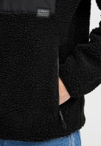 Black fleece pullover with a textured exterior, a zippered pocket, a smooth fabric patch, and a logo label on the chest area.