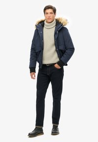 Doudoune marine avec capuche en fourrure, pull col roulé tricoté gris, jeans en denim foncé et bottes noires à lacets. Coupe décontractée avec poches.