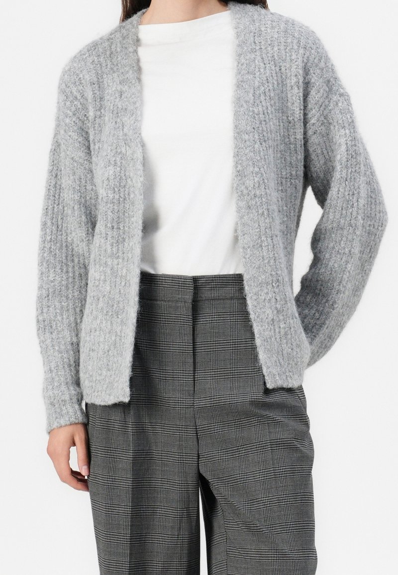 Femme portant un cardigan gris doux et texturé sur une chemise blanche et un pantalon gris à carreaux taille haute.