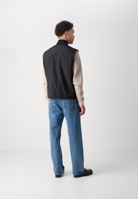 Schwarze ärmellose Weste mit hohem Kragen über einem beigen Langarmshirt, kombiniert mit hellblauen Jeans und schwarzen Schuhen.