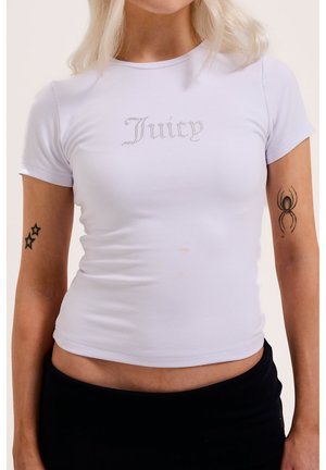 Frau trägt eng anliegendes weißes T-Shirt mit dem Schriftzug „Juicy“ aus Strasssteinen, zeigt Stern-Tattoos am linken Arm und ein Spinnen-Tattoo am rechten Arm.