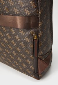 Sac à dos en tissu marron texturé avec un motif géométrique, doté d'une sangle en tissu marron uni et d'accents de fermeture éclair dorés sur le côté.
