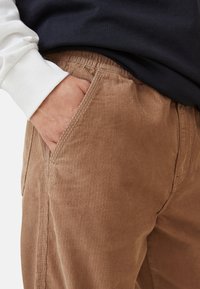 Person som bär bruna kordbyxorna med höger hand delvis i framfickan, klädd i vit skjorta och mörk topp.