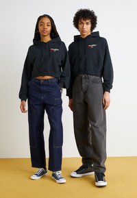 Svart avkortet hettegenser med frontlomme og blomsterbroderi, kombinert med lyse og mørke denimbukser samt kanvas-sneakers i svart og marineblå.