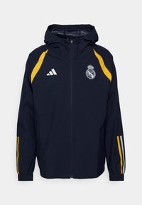 Donkerblauw gewatteerd jack met capuchon, gele strepen op schouders en mouwen, witte Adidas-logo en Real Madrid-embleem op de borst.