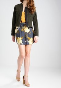 Femme portant une veste vert foncé sur un haut moutarde, une jupe bleu marine avec un imprimé floral jaune, et des sandales à talons beiges, debout sur fond blanc.