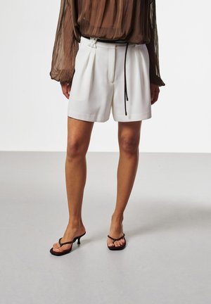 Person, die weiße, hoch taillierte, plissierte Shorts, eine braune durchsichtige Langarmbluse und schwarze Sandalen mit Absatz trägt und auf einem grauen Boden steht.