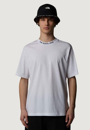 Giovane uomo che indossa una maglietta bianca The North Face con logo sulla scollatura e un cappello a secchiello nero The North Face su uno sfondo semplice.
