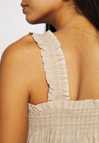Top in misto beige con arricciature, dotato di spalline volanti e di un tessuto testurizzato con un sottile motivo a quadretti. Spalle e schiena scoperte.